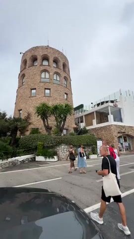 A video shows a cylindrical stone tower with arched windows and a white modern building with multiple levels behind it. Several people are walking on a paved road in front of these buildings. The man in the foreground is wearing a black t-shirt, black shorts, and white sneakers, and is carrying a white tote bag over his shoulder. He is looking to his right, towards the tower. A few other people are walking away from the camera, towards the modern building. Palm trees and greenery are visible around the tower and the modern building. The sky is overcast. A car is partially visible in the foreground on the left side of the frame.