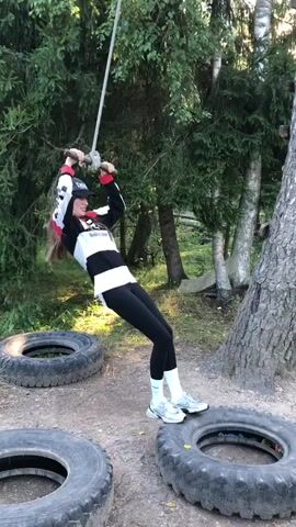 A young girl is in a forest, holding onto a rope swing attached to a tree. She is wearing a baseball cap, a long-sleeved shirt with black and white stripes, black leggings, and athletic shoes. She is standing on top of a large tire. In the background, there are more tires and trees. The girl is swinging on the rope.