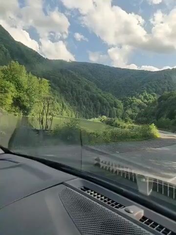 The video shows a view from inside a car driving along a road. The road curves to the right and is bordered by a body of water on the left and a lush green forest-covered mountain range on the right. The sky is blue with white, fluffy clouds. The car's dashboard is visible in the foreground.