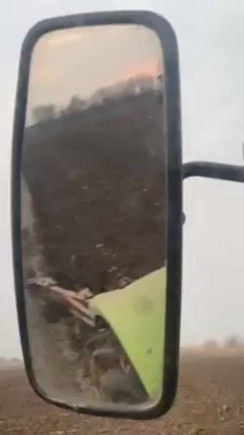 The video shows the reflection in a tractor's side mirror. The mirror reflects a plowed field with a green and red plow visible in the foreground. A line of trees is visible on the horizon, and the sky is overcast with a hint of pink or orange. The tractor is moving forward.
