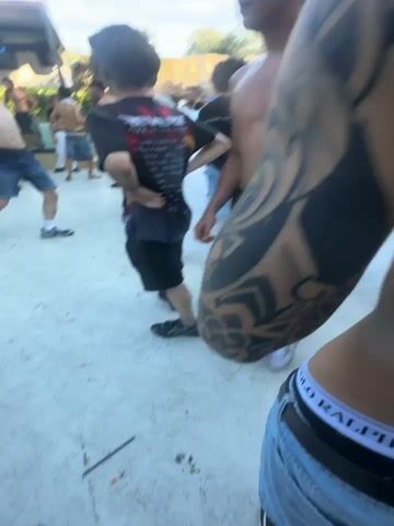 The video depicts an outdoor gathering. In the foreground, two individuals with prominent tattoos are visible, one from the chest up and the other from the waist up. Both are holding cans, suggesting they are consuming beverages. The camera is positioned close to their faces, creating a sense of intimacy or a first-person perspective. In the mid-ground, a group of people are mingling, with some interacting and others observing. White smoke or fog is present throughout the area, creating a hazy atmosphere. The background reveals a setting with natural elements, possibly a quarry or a rocky landscape, with trees and a bright sky overhead. The overall impression is of a relaxed, informal event, possibly a party or festival, with a focus on social interaction and enjoyment.