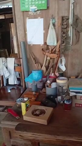 The video shows a workbench cluttered with various items. There are tools, containers, and what appear to be woodworking projects. A calendar hangs on the wall in the background. A close-up view reveals a wooden object with a circular indentation, possibly a mold or a base for a craft. Several cans and jars are also visible on the bench. The overall impression is of a workspace that is actively used, with materials and tools readily accessible.