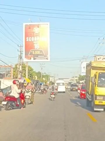 The video shows a street scene with moderate traffic under a clear sky. A large billboard advertising "RED SCORPION" and "16" with a price of "9.000" is prominently displayed above the road. The billboard is illuminated by the sun, making some details difficult to discern. A variety of vehicles, including motorcycles, cars, and a truck, are visible on the road. Several people are riding motorcycles, some wearing helmets and face coverings. On the sides of the road, there are stalls or small shops, with some people gathered around them. Power lines crisscross the sky, and utility poles line the street. The overall impression is of a busy, sun-drenched urban or semi-urban environment.