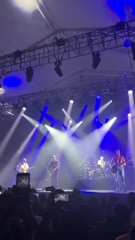 The video shows a band of five musicians performing on a stage under a large tent. Four members are playing guitars, while one is on the drums. Bright white and blue stage lights illuminate the band. A crowd of people is visible in the foreground, many holding up their phones to record the performance. The stage is set up with a complex overhead lighting and support structure.