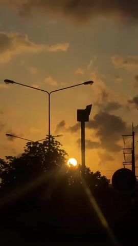 The video shows a time-lapse of a sunset. Silhouetted against the warm orange sky are two streetlights, one taller than the other, with their lamps angled downwards. In the foreground, the dark silhouette of a dense tree obscures the lower half of the frame, with sunlight peeking through its branches. As the time-lapse progresses, the sun, a bright orb, becomes more visible and eventually rises higher in the sky, its rays casting diagonal lines across the scene. The sky is filled with scattered clouds, tinged with orange and yellow from the sunset. In the background, hints of distant trees and possibly power lines can be seen. The overall impression is a peaceful transition from sunset to evening.