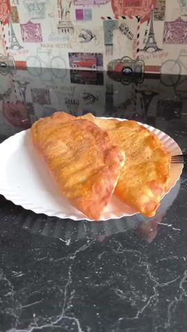 The video features two golden-brown, freshly fried pastries, possibly chebureki, displayed on a white, ridged paper plate. The plate rests on a dark, speckled countertop. In the background, there is a light-colored wallpaper or print with vintage travel-themed illustrations and text. Visible text on the wallpaper includes "PARK-HOTEL", "BON VOYAGE", and "PARIS". The illustrations include a red vintage car, bicycles, and the Eiffel Tower. The camera slowly pans across the pastries, providing a close-up view. No people are visible in the scene.