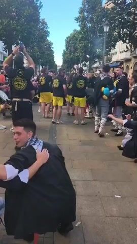 A group of people in black t-shirts with yellow emblems and yellow shorts are playing brass instruments in a street. Some people in the foreground are wearing black robes. There are trees and buildings in the background. The ground is paved with bricks. Some people are holding up their phones to record the event. The sky is blue.