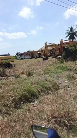 The video shows a line of yellow excavators parked on a grassy, unkempt lot. The excavators are stationary, and their arms are raised or lowered, suggesting they are not in use. The lot appears to be overgrown with dry grass and weeds. In the background, there is a building with a corrugated metal roof, and a few trees, including a palm tree. The sky is blue with some clouds. The video is filmed from a moving vehicle, possibly a motorcycle, as indicated by the partial view of a mirror and handlebars at the bottom of the frame. The overall impression is of an abandoned or disused construction equipment lot.