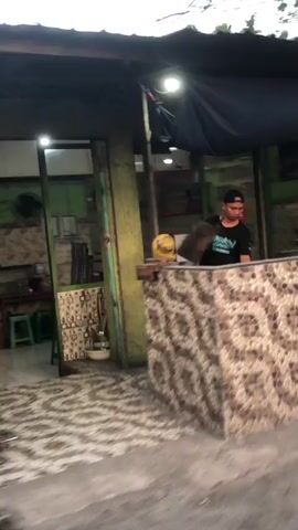 The media file shows a man standing behind a counter in what appears to be an outdoor eatery or shop. The man is wearing a black t-shirt with a graphic print and a black baseball cap. He is looking towards the viewer. The counter is decorated with a mosaic tile pattern. Behind the counter, there are shelves with various items, and a green wall is visible. To the left, there is an open doorway leading into what looks like a seating area with green stools. The floor is paved with a patterned tile and the ground in front is a rough concrete surface. A yellow object, possibly a ball or a piece of fruit, rests on the counter. The lighting suggests it is either early morning or late afternoon, with soft, natural light illuminating the scene. The overall atmosphere is casual and everyday. The location cues, such as the architecture and the general appearance of the establishment, are consistent with a setting in Kejapanan, Indonesia. There is no discernible text visible in the media file.