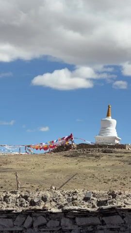 The video captures a serene daytime landscape featuring a prominent white Buddhist stupa with a golden spire, situated on a dry, earthy hill. Colorful prayer flags, strung between poles, extend across the middle ground, fluttering gently in the wind. The sky is a vibrant blue, adorned with scattered white clouds that drift slowly. A low stone wall made of rough-hewn rocks is visible in the foreground. The camera slowly pans from right to left, revealing more of the arid terrain and the full span of the prayer flags. No people are visible, and there is no discernible text. The scene suggests a peaceful, high-altitude environment, consistent with Labrang township, China.