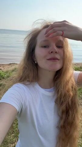 A young woman with long, wavy, reddish-blonde hair is captured in a selfie video. She is outdoors, standing on grassy ground with a body of water and a sandy shore in the background. The sky above is blue with white clouds, suggesting it is daytime and fair weather. The woman is wearing a white t-shirt and has a pearl earring in her visible ear. She is smiling and touching her hair, appearing relaxed and happy. The setting appears to be a natural environment, possibly a park or a beach, given the presence of grass, sand, and water. There is no visible text in the video. The overall mood is cheerful and carefree.