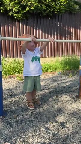 A toddler, wearing a white t-shirt, green shorts, and brown sandals, is hanging from a metal bar. The child also has a brown hat. The playground is covered in gravel. There is a brown fence and some greenery in the background.