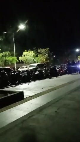 The video shows a dimly lit outdoor scene at night. Several motorcycles are parked in a row on the left side of the frame. Beyond the motorcycles, there are several cars parked. There are trees and streetlights visible in the background, illuminating the area with a yellowish glow. A sidewalk or paved area is in the foreground. A few people are visible standing and walking in the background. The overall impression is of a parking area at night.