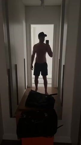 A man takes a selfie in a hallway mirror. He is shirtless and wearing black shorts and a black baseball cap. There are two large duffel bags on the floor in front of him, and he is holding a smartphone in his right hand, taking the photo. The hallway has white walls and closets. The lighting changes from dim to bright, highlighting the man and the bags.