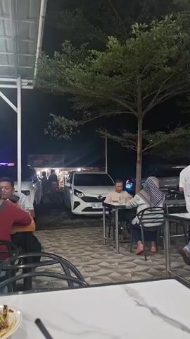 The video shows a nighttime outdoor scene at what appears to be a food stall or market. In the foreground, there are tables and chairs, with a portion of a white table with a marbled pattern visible. In the middle ground, there are several people seated at tables. On the left, a person wearing a red top is seated at a table. In the center, a man wearing a light-colored shirt is seated at a table, facing a woman wearing a headscarf and a striped top. They are both looking down at their table. Behind them, several cars are parked, including a white car with its headlights on. In the background, there are more stalls or shops with lights. A large tree with green foliage is prominent in the middle of the frame, casting shadows. The overall lighting suggests it is dark outside.