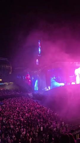 The video captures a night concert with a large crowd in an amphitheater. The stage is elaborately lit with pink, purple, and blue lights, and features a large screen displaying visuals. There are pyrotechnics and fireworks going off above the stage, adding to the spectacle. The audience is densely packed and appears to be actively engaged with the performance. The camera is positioned from an elevated vantage point, looking down on the crowd and the stage.