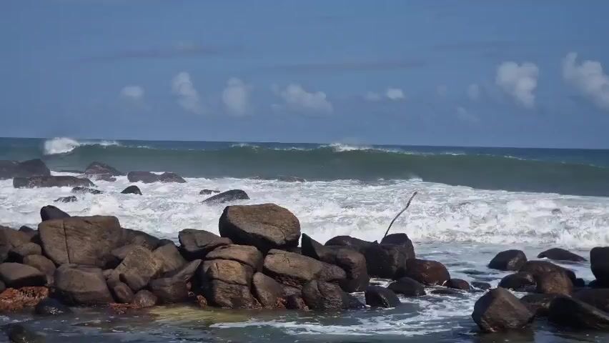 The video depicts a rocky coastline under a blue sky dotted with puffy white clouds.  Large, dark gray rocks form a barrier near the foreground, partially submerged in the ocean water. Behind the rocks, waves crash and foam, creating whitecaps as they approach the shore. The sea appears a greenish-blue in the distance. A thin, dried branch is stuck in the rocks. The waves are continuous, indicating a moderately rough sea condition.