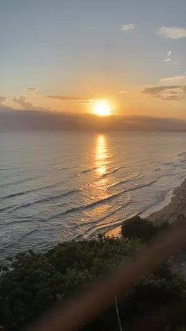 This is a vertical, high-angle shot of a coastal sunset. The sun, a bright orange disc, is low on the horizon, casting a shimmering, golden reflection across the calm sea. Gentle waves roll towards a sandy beach lined with trees and vegetation. The sky transitions from a pale blue overhead to soft orange and yellow hues near the horizon, with a few wispy clouds. A wooden railing is visible in the foreground, partially obscuring the view. There are no people or discernible man-made objects in the water or on the beach. The overall impression is one of natural tranquility and scenic beauty.