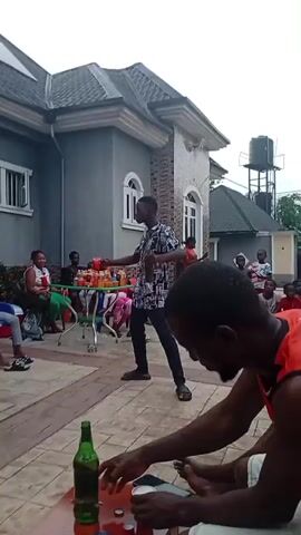 The video shows an outdoor gathering of people in what appears to be a residential area in Onicha, Nigeria. The scene is set on a paved courtyard or patio with a large grey house featuring white trim in the background, and another structure with a dark roof and a water tank on a stand further behind.

The central figure is a man wearing a patterned short-sleeved shirt, dark trousers, and sandals. He is actively dancing or performing, holding a dark bottle in one hand and a red plastic cup in the other. He moves rhythmically, engaging with the audience.

Around him, various people, including adults and numerous children, are gathered. Many are seated on colorful plastic chairs or directly on the ground, observing the man. Some women are also visible, sitting on chairs. In the middle ground, there is a small rolling cart or table laden with several red plastic cups, presumably filled with drinks, suggesting a social event or celebration.

In the immediate foreground, part of another person is visible, specifically their arms, as they are in the process of opening a green glass bottle on the ground, next to another identical bottle that has already been opened.

The lighting indicates it is daytime, and the overall atmosphere appears relaxed and convivial, with people enjoying the performance. There is no discernible text visible on any objects or clothing in the video.