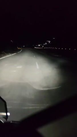 The video shows a first-person view of someone riding a motorcycle at night on a paved road. The headlight beam illuminates the road ahead, revealing white lane markings and some scattered debris. The surrounding environment is dark, with faint lights visible in the distance along the side of the road, suggesting a populated area or town. The rider's hands and part of the motorcycle's handlebars and mirrors are visible in the foreground. The road appears to be winding, and there is a slight incline in the distance. The headlight beam casts a hazy glow, indicating the presence of some moisture or dust in the air.