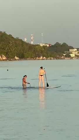 Two people are in the water near a coastline. One person, standing in the water, appears to be holding a rope attached to a paddleboard. The other person, on the paddleboard, is bending over. The water is calm and reflects the sky. In the background, a green, tree-covered hill with buildings and towers is visible. The sky is a clear, pale blue.