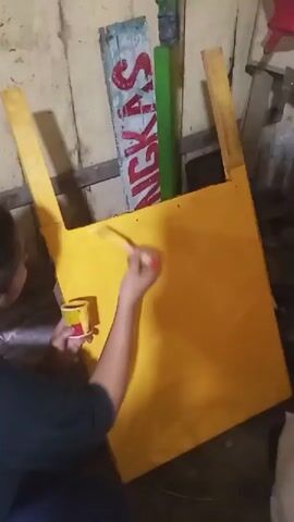 A person is shown painting a yellow object with a red and white brush. The person is holding a small yellow container of paint in their left hand. The yellow object appears to be a large, flat surface with some supporting structures. Behind the yellow object, there are two vertical planks of wood, one with red lettering that appears to say "NGKAS", and another that is green. The environment appears to be a workshop or storage area with wooden walls.