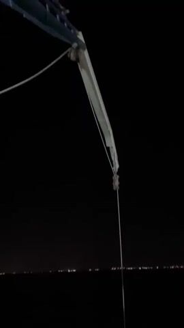 A close-up, low-angle shot shows a long metal pole extending from a white crane mechanism down into murky green water at night. The water is illuminated by an unseen light source, causing it to glow. In the background, a distant, blurred line of city lights is visible against the dark night sky. The pole enters the water, creating a disturbance with some foam and bubbles.