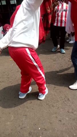 This video appears to capture a moment of collective physical activity, possibly a school event or a community gathering in Sukaraja, Indonesia. The subjects are primarily people, many of whom are dressed in matching red and white attire, suggesting a uniform or team outfit. Some individuals are wearing red shirts with white accents, red pants with white stripes, and white shirts. Others are in red tops and dark pants, or in blue jeans. The setting is an outdoor, paved area, likely a schoolyard or public space, with some greenery and buildings visible in the background. The lighting suggests it's daytime, possibly morning or afternoon, with shadows indicating the sun is out, so the weather is likely fair.

The activity seems to involve synchronized movement or exercise. In the foreground, a person in a white shirt and red pants is in a push-up or plank position, with their legs extended. Another person, also in red and white, is in a similar pose. The overall impression is one of order and participation in a group activity. There are hints of community interaction, with some individuals holding hands or standing close together. No discernible text is visible in the video.