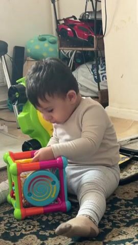 The video shows a baby sitting on a carpeted floor, playing with a colorful, multi-sided activity cube. The baby is wearing a light-colored long-sleeved shirt and grey pants with a striped pattern. The activity cube is bright and has various textures and interactive elements. The baby is leaning forward, with their face resting on top of the cube and their hands touching it. In the background, there are shelves with toys, including a red car and a green toy lawnmower with a Batman logo. The environment appears to be a living room or play area, with some clutter visible. The lighting is ambient and natural.