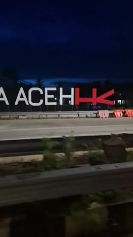 The video displays a large white sign with the text "SIGLI - BANDA ACEH" in a sans-serif font, illuminated at night. The last two letters "I K" are in red. The sign is positioned above a road or highway, with a dark blue night sky in the background. In the foreground, there are orange and white traffic barriers or construction barriers. The camera pans from left to right, revealing more of the sign and the road. The ground is dark asphalt. There are some faint light sources, possibly from distant vehicles or streetlights, visible in the background and on the road.