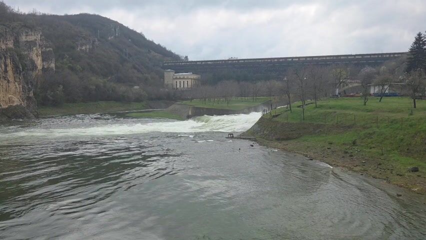 The scene presents a water reservoir or dam in a landscape setting. The river flows strongly into the foreground, with turbulent water creating white foam near the edge of a small concrete structure resembling a spillway. Behind the spillway is a building next to a line of trees. The dam, a large gray structure, is visible in the background along with green hills and a cloudy sky. The river banks are covered with green grass. The scene is tranquil but shows signs of water management and human impact on the natural environment.