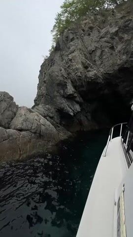 The video shows a boat navigating through a narrow passage between rocky cliffs. The water is dark but slightly translucent, with a hint of green. The cliffs are gray and rugged, with a cave entrance visible on the right. The sky is overcast, adding to the somber atmosphere. The camera is positioned on the boat, providing a view along the side and towards the front. The boat's white exterior is visible in the foreground. The overall scene is reminiscent of a secluded coastal area.