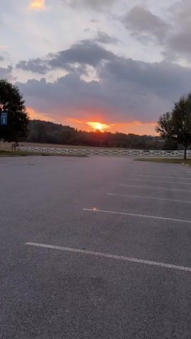 The video shows a sunset over a rural landscape. The foreground is a dark asphalt parking lot with white painted parking lines. In the middle ground, there is a white picket fence and a line of trees. The background features rolling hills covered in trees, with the sun setting behind them. The sky is a mix of orange, yellow, and grey clouds. A lamppost with two lamps is visible on the right side of the frame, near some trees and grass. The lighting suggests it is either early morning or late evening.