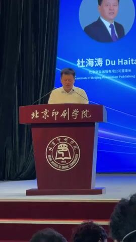 A man stands behind a dark red podium. He is wearing a white polo shirt and appears to be speaking. The podium has Chinese characters and a circular emblem on it. Behind the man is a large blue screen with a smaller circular image of another man in the upper right corner. The text on the screen is in both Chinese and English. The man on the screen is identified as Du Haitao, Chairman of Beijing Promunion Publishing Co., Ltd. The environment appears to be an indoor event, possibly a conference or presentation, with dark curtains in the background and an audience visible in the foreground.
