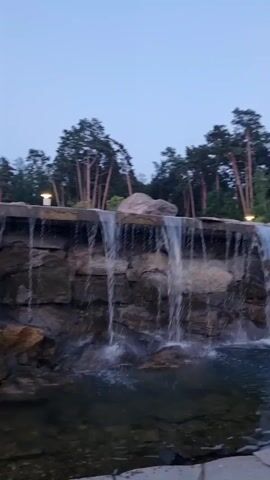 A man-made waterfall cascades over a rocky embankment into a pool of water. Tall, slender trees, likely pines, with sparse foliage, form a dense forest in the background. The sky is a pale, overcast blue, suggesting it is either early morning or late evening. The overall scene is serene and natural, with the man-made waterfall blending seamlessly into the surrounding environment.