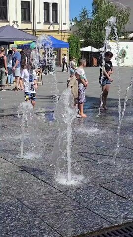 A group of children and adults are gathered in an outdoor public space. The foreground is dominated by a water feature with several jets of water spraying upwards from the pavement. Several children are interacting with the water jets, splashing and playing. One child in the center is being held by an adult. In the background, there are buildings, tents, and more people. The overall scene appears to be a sunny day in a town square or park.