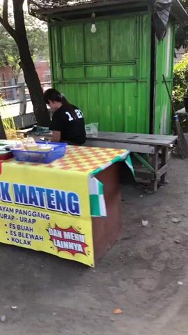 A person wearing a black t-shirt with the number 12 on the back is sitting on a wooden bench in front of a green food stall. The stall has a yellow banner with blue and red text that reads "AYAM PANGGANG URAP - URAP ES BUAH ES BLEWAH KOLAK DAN MENU LAINNYA". There are various food items displayed on the counter. The ground is dirt and gravel. A tree is visible to the left of the stall, and a building with a red roof is in the background. The lighting suggests it is daytime.
