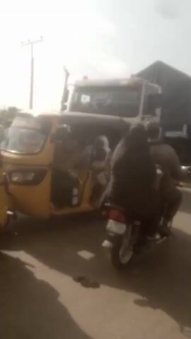 The video shows a street scene in what appears to be a congested area. Several large trucks are present, some covered with tarpaulins. A motorcycle carrying two individuals is maneuvering through the traffic, driving to the left of the screen. Other motorcycles and vehicles can be seen in the background. The weather appears bright and sunny.
