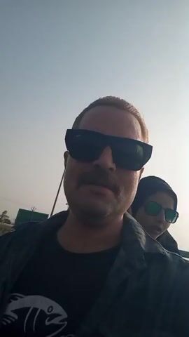 A man in dark sunglasses and a plaid shirt over a black t-shirt is taking a selfie video outside. He has a mustache and short blond hair. Behind him is a woman in sunglasses and a head covering. They appear to be stopped or moving slowly on a road with trucks and buildings in the background. A light pole is visible. The sky is bright and hazy.