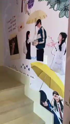 The video shows a staircase decorated with large illustrations of Korean dramas. The illustrations on the wall depict scenes from various dramas, including couples under umbrellas, school scenes, and romantic images. Text in Korean and English is incorporated into the artwork. The stairs are made of a light-colored material, and the wall is painted white.