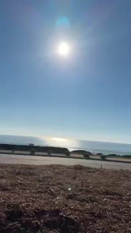 The video presents a sunlit coastal view, with the bright sun high in a clear blue sky, reflecting intensely on the ocean's surface. The camera appears to be moving, possibly from a vehicle traveling along a road near the coast. The immediate foreground features ground cover consisting of wood chips or mulch, transitioning into shrubbery and sparse vegetation. A glimpse of a guardrail separates the foreground from the ocean view. There are also what appear to be rail tracks in the foreground. The scene conveys a sunny, coastal landscape.