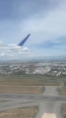 The video captures an aerial view from an airplane window, showing the wing of the plane with a blue accent and a sprawling cityscape beneath a partly cloudy sky. The runway is visible below, indicating the plane is approaching for landing. The landscape is a mix of urban development with buildings and infrastructure and some green spaces. The overall impression is a descent into a densely populated urban area.