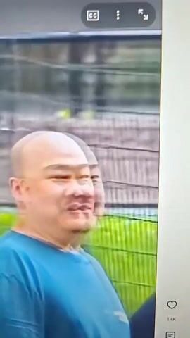 A group of people are walking in what appears to be a park. The first shot focuses on a bald man in a blue shirt, who turns to speak to a woman laughing. The second shot shows the bald man talking to the woman, and then the perspective shifts to show a group of men walking in the park. One man is wearing a blue shirt and dark shorts and carrying a backpack, another is in a light blue shirt, and a third is in a dark blue shirt. A fourth man in a dark blue shirt and dark shorts is also visible. The background consists of trees and greenery.