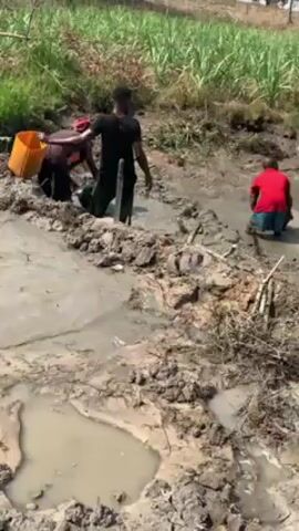 The video shows three people working in a muddy field. One person is wearing a red shirt and blue shorts, kneeling in the mud and using a black container to move mud. Another person, wearing a dark shirt and long pants, is standing in the mud while the third person, wearing a head covering and dark clothing, carries a yellow bucket through the mud. The field is uneven and has several puddles. In the background, there is tall green vegetation. The overall impression is of manual labor being performed in a wet and muddy environment.