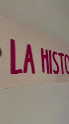A close-up shot pans across a light-pink sign with raised, dark pink lettering that reads "HISTORIA DEL CAFÉ". The letters are slightly blurred due to motion. The sign is mounted on a textured, light-colored wall. Below the sign, there is a green painted trim. The footage then shifts to show three framed black and white illustrations or comic panels hanging on the wall. The leftmost panel is partially visible. The middle panel depicts a cartoonish scene with stacked coins and a speech bubble. The rightmost panel shows a cartoonish depiction of a bus or vehicle with a smiling face. The illustrations are within a green frame.