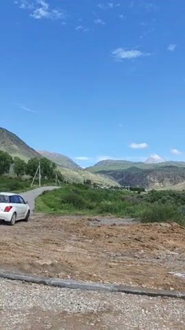 The video shows a scenic landscape under a bright blue sky with scattered white clouds. In the foreground, there is a patch of loose dirt with a gravel and curb border. To the left, a white car is parked on the side of a paved road that curves into the distance. The road leads to a valley with rolling green hills and mountains. The hills are covered in lush green grass and some scattered bushes. In the middle ground, there are small buildings visible, suggesting a rural settlement. The overall impression is one of a peaceful, natural environment. The video pans slightly to the right, offering a wider view of the valley and mountains.