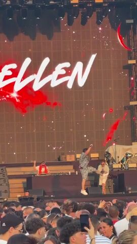 A concert is taking place on a stage with a large screen displaying the word "QUELLEN" in white text against a reddish, abstract background. There are several people on stage. In the center, a person in a patterned shirt and shorts is dancing, kicking their leg in the air. To the left, a person wearing sunglasses and a red cloth around their neck stands behind a DJ booth. To the right, another person is kneeling. In front of the stage, a large crowd of people are watching the performance, many holding up their phones to record. The environment appears to be outdoors, likely during the day, given the lighting.