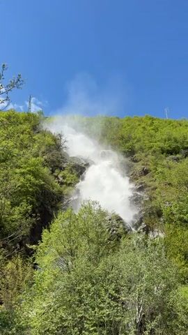 The video shows a waterfall cascading down a steep, green hillside under a clear blue sky. The waterfall is the main focus, appearing white and powerful as it descends over multiple tiers of rock and vegetation. Lush green trees and bushes cover the hillside around the waterfall. Two electric power poles are visible at the top of the hill.