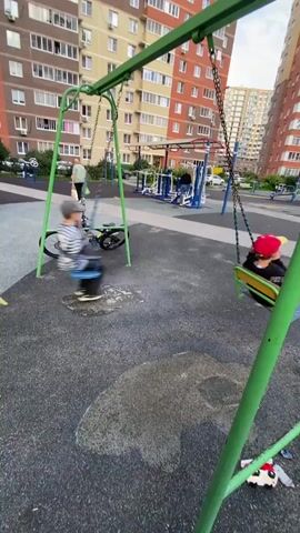 The video shows two children on swings on a playground. In the foreground, a child wearing a red hat and a black and white shirt is swinging. In the background, another child wearing a striped shirt and a hat is also swinging. A bicycle with a yellow frame and black tires is parked near the swings. In the background, there are apartment buildings and other playground equipment. The ground is covered with a dark, speckled surface. The video is recorded from a low angle.