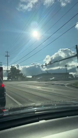 A vertical video shows a view from inside a car at an intersection with traffic lights. The sun is shining brightly in a blue sky with some white clouds. There are power lines stretching across the sky. In the distance, there are buildings and trees. Cars are visible on the road and crossing the intersection. The video is taken from a stationary viewpoint, implying the car is stopped at the intersection. The overall scene is a common urban or suburban street view.